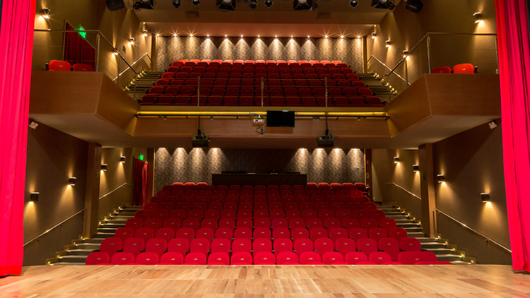 Uma visão da plateia do Teatro Cesgranrio, com poltronas vermelhas, de cima do palco, margeado por cortinas também vermelhas