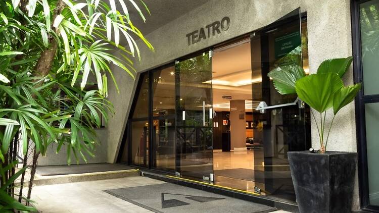 Entrada do teatro com portas de vidro escuro, um tapete e vasos de plantas