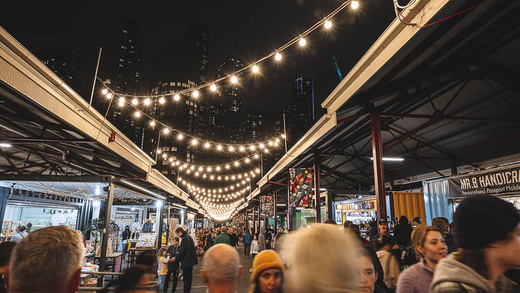 Strings of lights are above people walking through a market. 