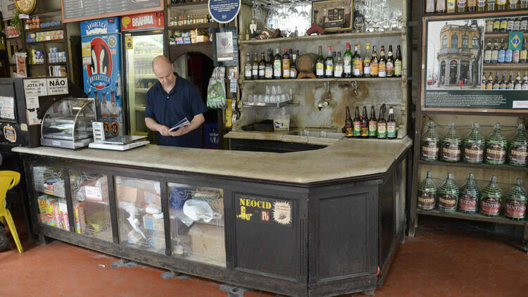 Um homem atrás de um balcão de bar, em frente à prateleiras com garrafas