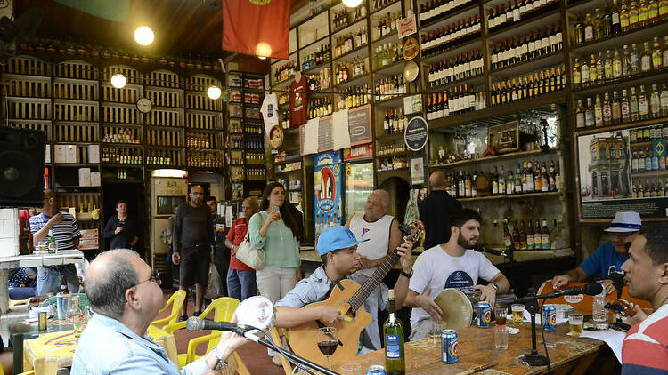 Cinco músicos se apresentam em uma roda de samba em um bar com prateleiras altas e abarrotadas de garrafas de bebidas