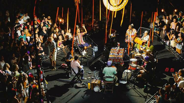 Uma roda de samba de apresenta em um palco hexagonal com o público ao seu redor e faixas coloridas pendem do teto