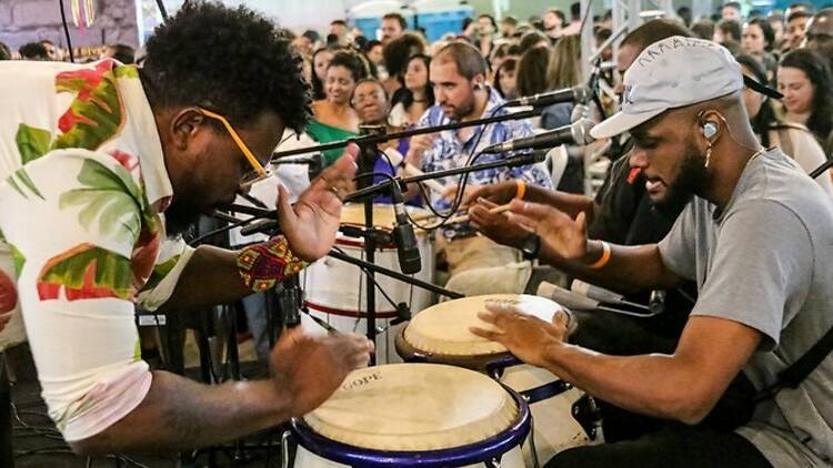 Dois mpusicos em destaque tocam em uma roda de samba com o público ao fundo