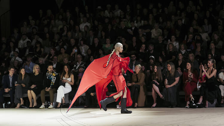 A model wearing a red garment walks on the runway. 