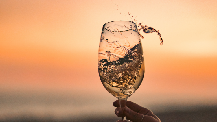 a hand swirling wine in a glass against a sunset backdrop
