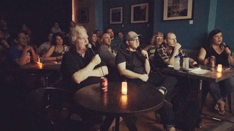 Comedians sit at tables in QED Astoria, holding microphones.