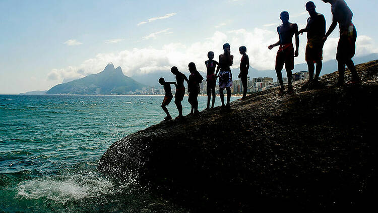 Silhueta de pessoas na Pedra do Arpoador pulando na água com o Morro Dois Irmãos ao fundo