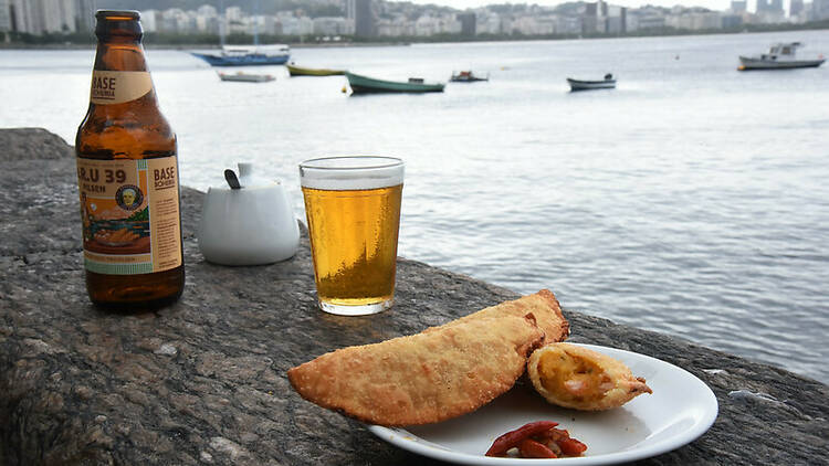 Uma garrafa de cerveja, um copo americano cheio e um prato com dois pastéis na Mureta da Urca