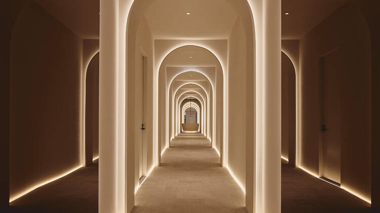 A long hallway lined with beige stone walls and flooring and ambient lighting. 