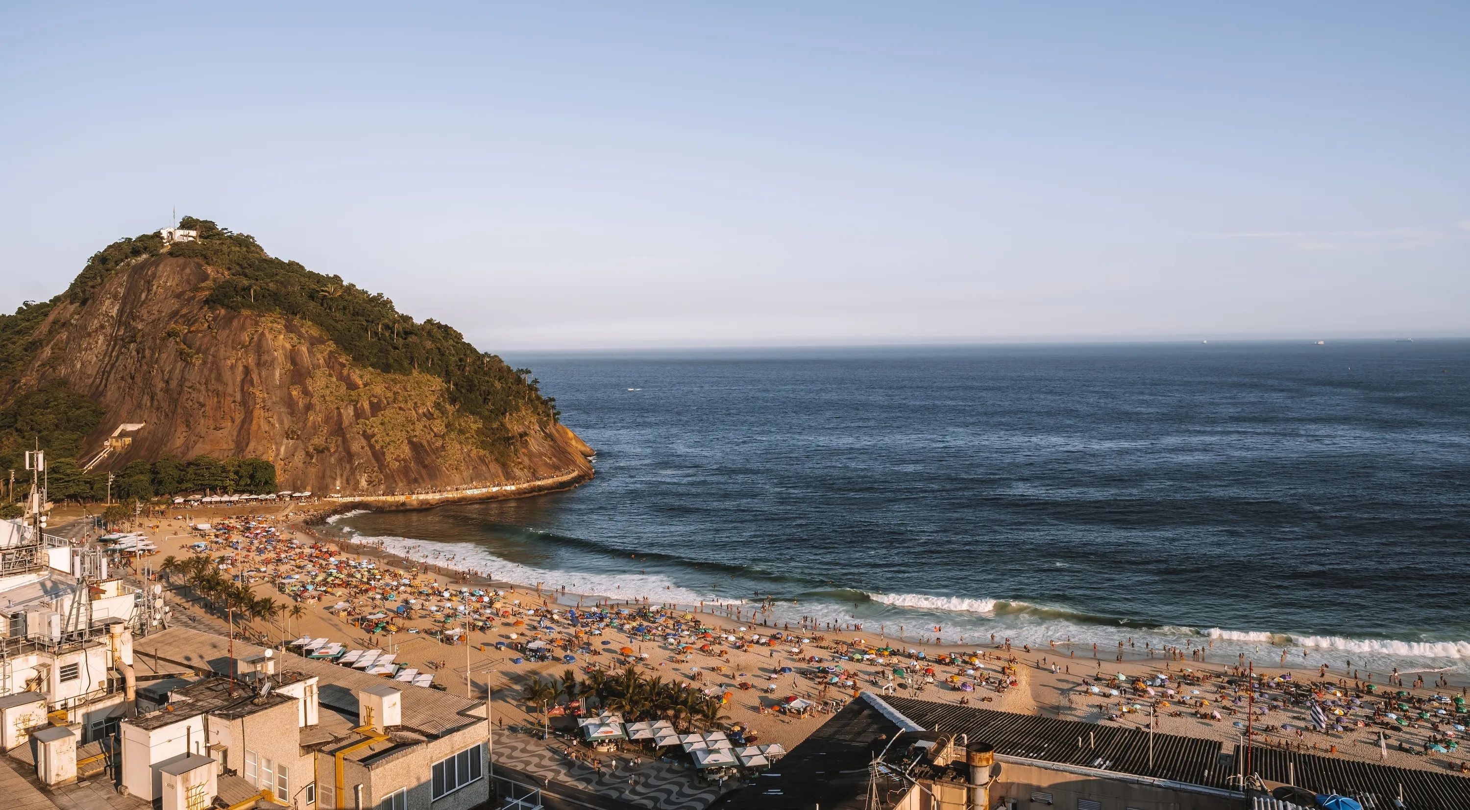 Faixa de areia da Praia do Leme cheia de banhistas, com mar esverdeado ao fundo e Pedra do Leme à esquerda