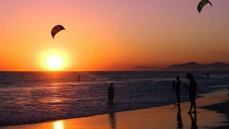 Pôr do sol na Praia do Pepê com silhuetas de parapentes e pessoas na água