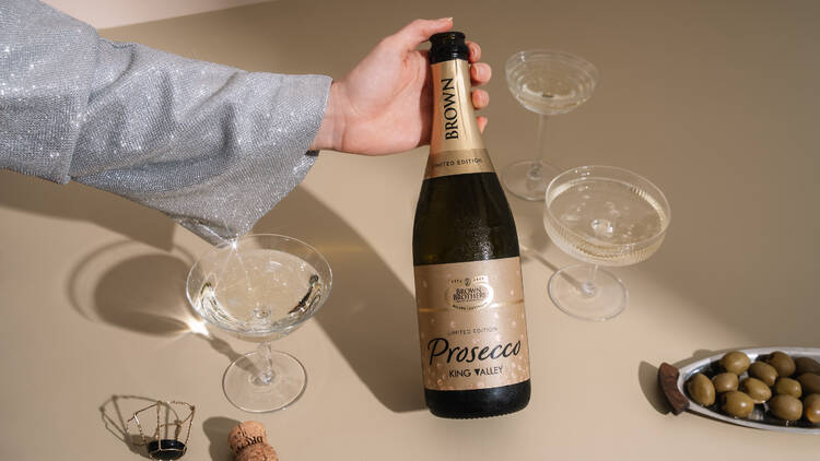 A person holding a bottle of Brown Brothers prosecco surrounded by glasses and a plate of olives.