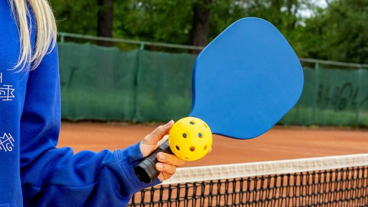 A woman with blonde hair holding a yellow ball and blue paddle on a pickleball court.