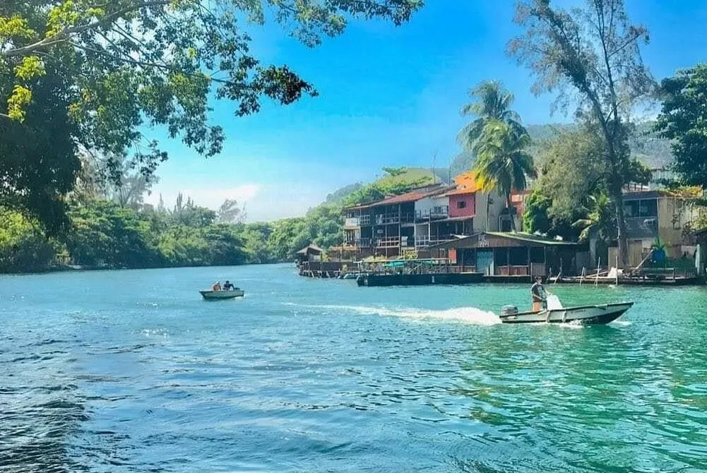 Dois barcos se locomovem na água com casas pequenas ao fundo