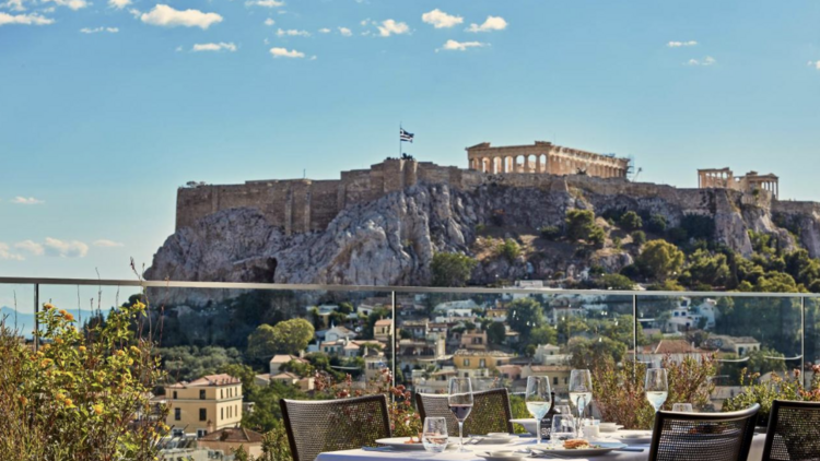 A hotel balcony with dining table and chairs at Electra Metropolis hotel in Athens, offering a captivating view of the Acropolis.