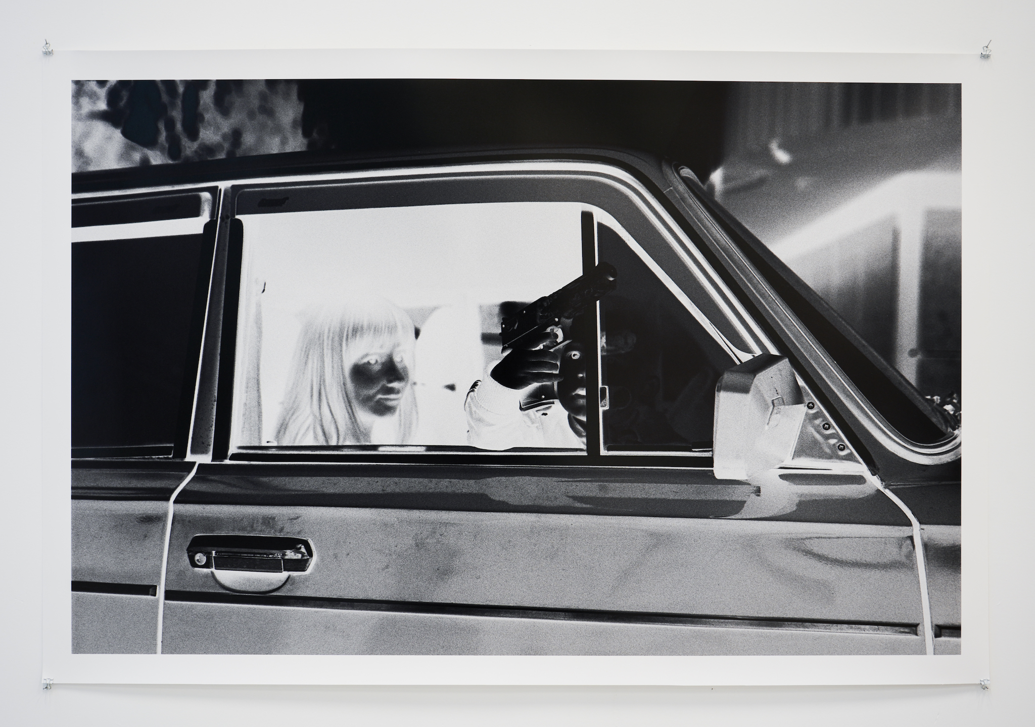 Two people sit in a car in a black-and-white photo. One holds a gun out the window.