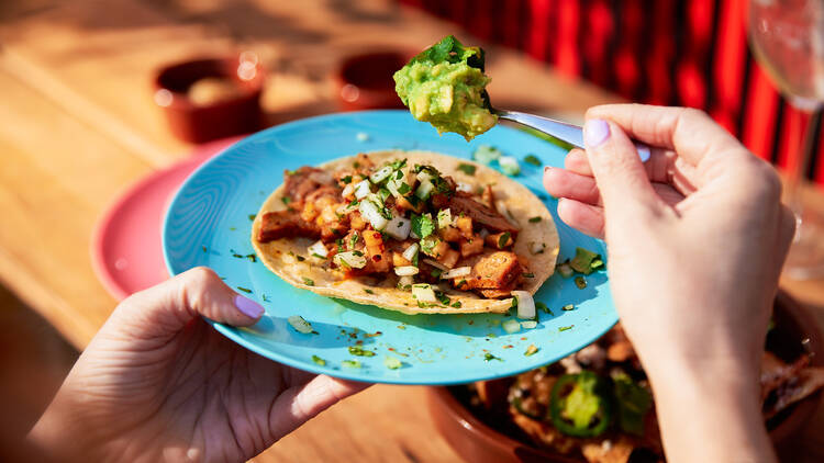 Person spooning guacamole into an open taco on a blue plate. 