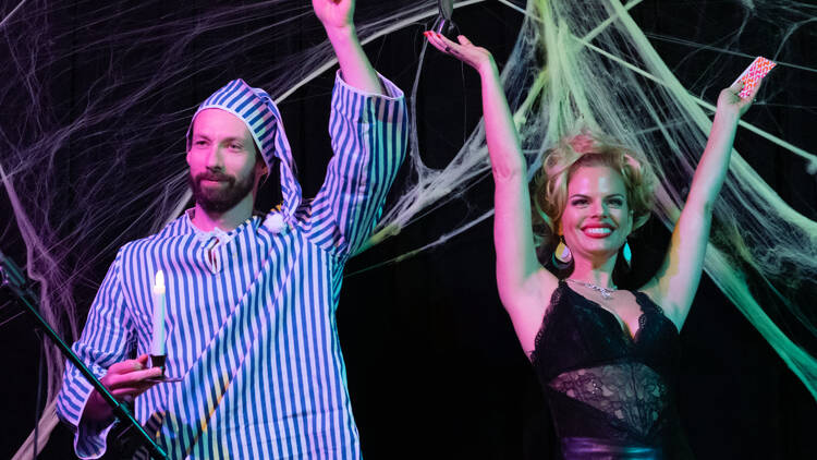 Two performers raise their hands in front of a wall of cobwebs.