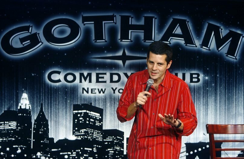 A man in a red shirt performs comedy on stage.