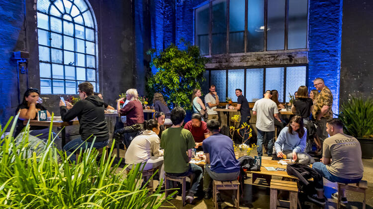 The Greenhouse Bar pop-up interior with lush greenery and people gathered around table with drinks.