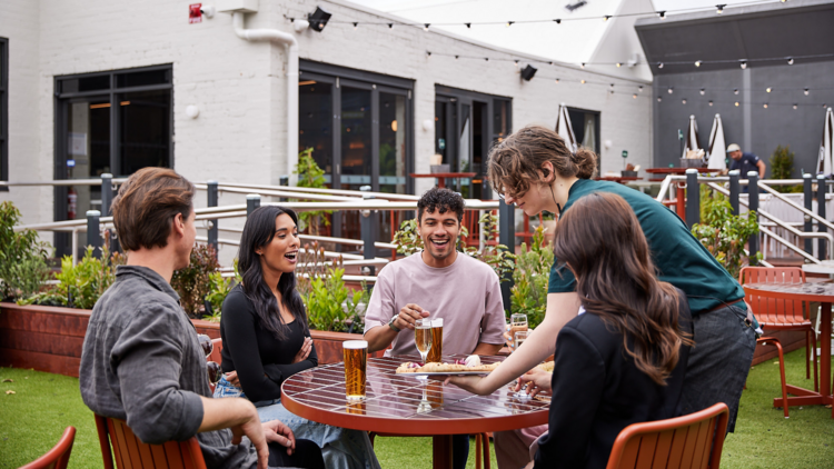 A staff member setting food down for a table of four, who are smiling in a beer garden