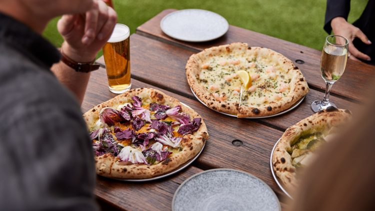 Three woodfired pizzas seen from above next to a pot of beer and a glass of sparkling wine