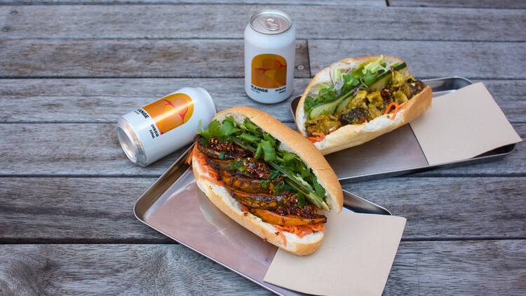 Assorted banh mi with canned beers and napkins on a wooden bench.