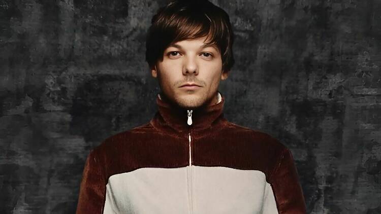 Louis Tomlinson veste casaco vermelho e branco e olha para a câmera