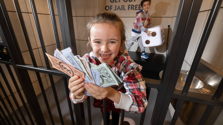 A child in a jail cell holding fake money at the Monopoly Dreams theme park.