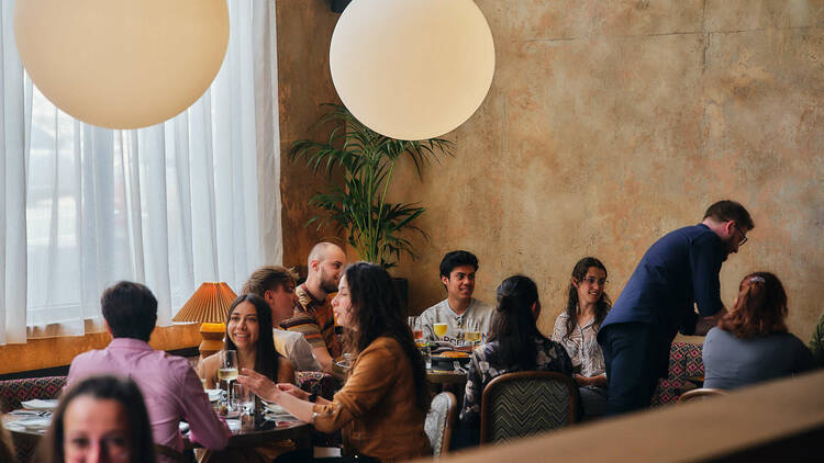 Tables of groups in a restaurant with spherical hanging lamps and greenery.