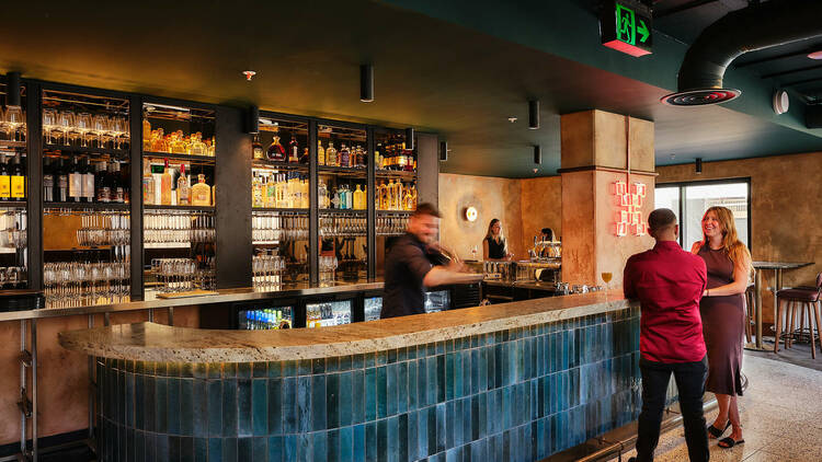 A  bartender shaking drinks behind a teal hue-tiled bar for two customers.