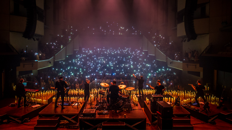 A rock band seen from the back of the stage, looking out onto a sea of candles and phone lights