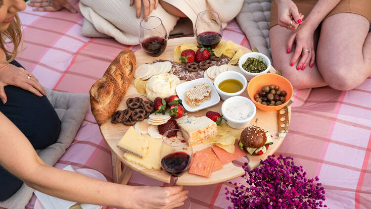 People on a picnic rug gathered around a small picnic table of food and wine.