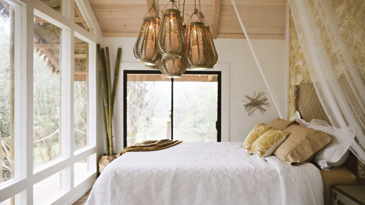 A bedroom, complete with a double bed, large floor to ceiling windows, and a statement chandelier, in a tropical treehouse in Fern Forest, Hawaii