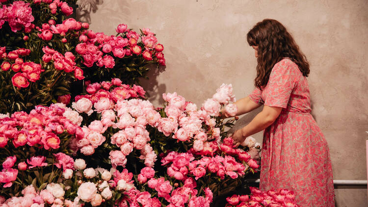 A person wearing a pink dress standing next to a huge pile of pink peony flowers. 