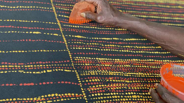 A close-up of a canvas with a black background, and yellow and red dots. A hand is holding a utensil and a tub of paint. 