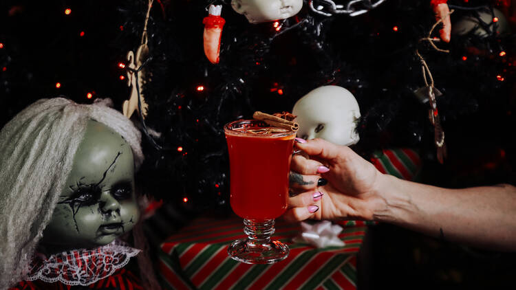 A woman holds up a drink with a creepy background consisting of doll heads.