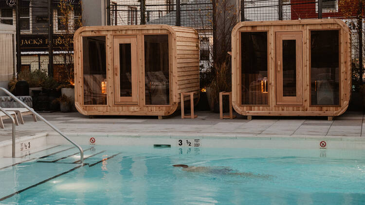 A person swims in an outdoor pool next to cedar saunas.