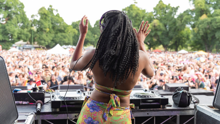 A DJ with long black coily hair stands behind the decks facing a Midsumma crowd