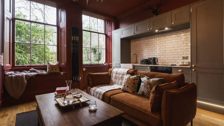 Living room with deep red walls, large bay windows, coffee table and a copper coloured sofa. 
