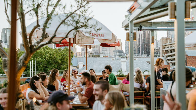 Busy rooftop bar with umbrellas and groups of people sitting at tables at dusk.. 