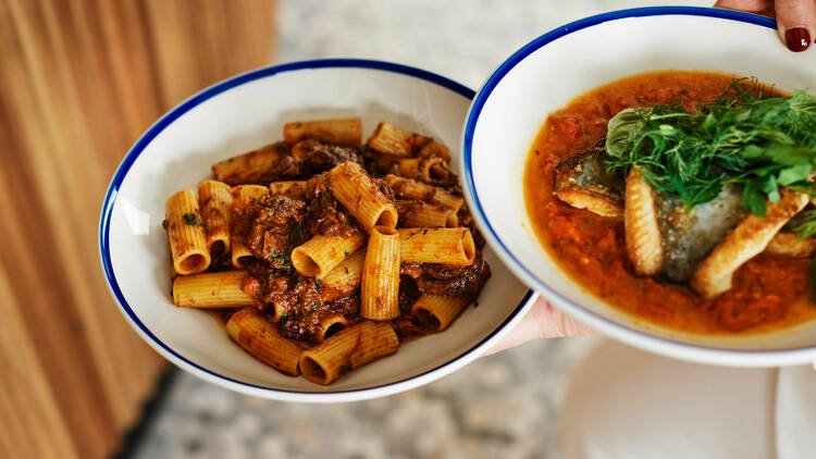 Woman holding a plate of beef cheek ragu and a plate of fish and greens.