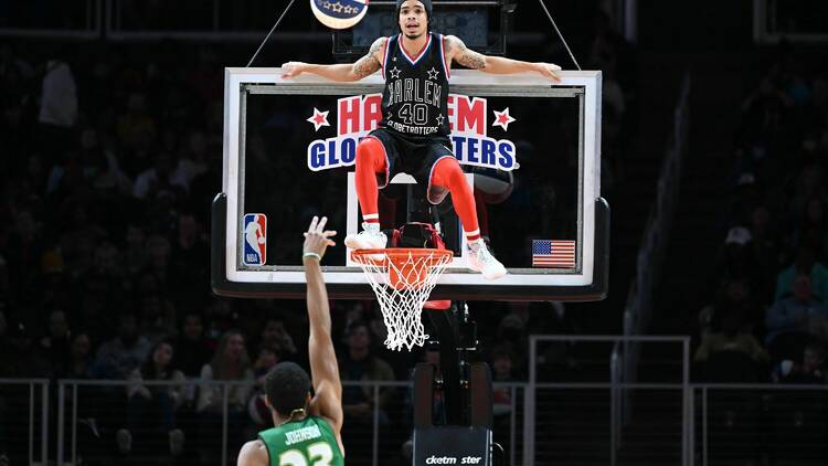A man stands on top of a basketball net while another shoots a basketball
