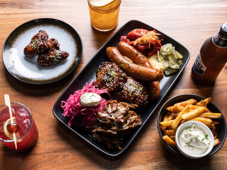 Assorted barbecue meats and sides with beers and cocktails on a wooden table.