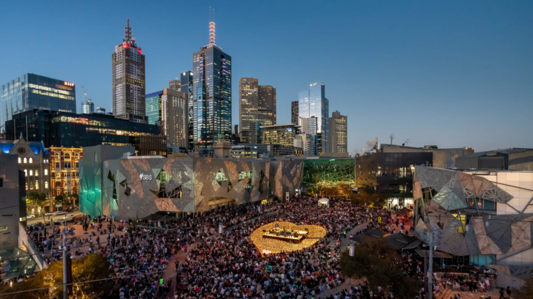 A stage surrounded by hundreds of glowing candles shaped as a heart, in Fed Square in front of the city skyline