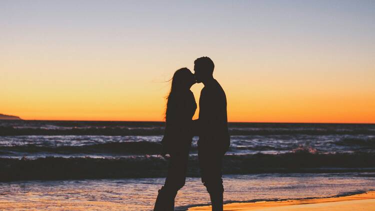 A couple kissing, silhouetted in front of the ocean at sunset. 