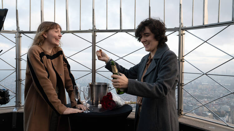A couple pops a bottle of champagne at the Empire State Building.