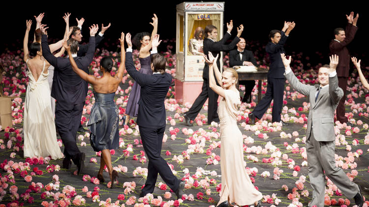 Dancers in a circular formation raising their hands above their heads. The stage is covered with red and pink flowers.