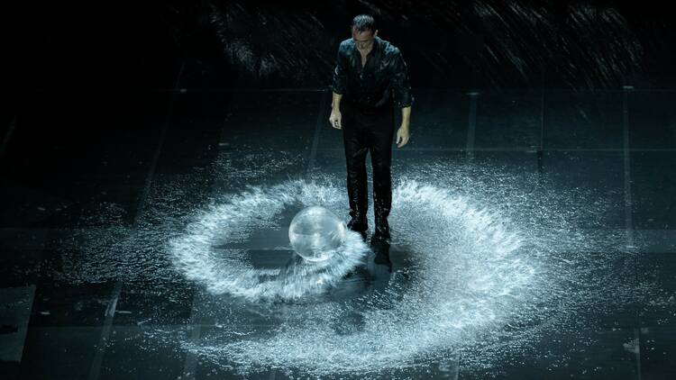 A man stands on a dark stage surrounded by a whirlpool of water