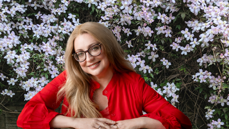 Van Badham wears a red shirt and glasses and appears in front of a garden wall covered in light pink flowers